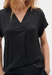 Blouse noire à manches courtes avec un col en V, texture légèrement froissée et coupe décontractée, présentant un drapé subtil à l'ourlet.