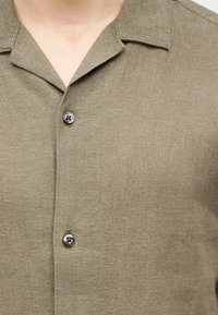 Ανδρικό πουκάμισο χακί με γιακά, δύο ορατά κουμπιά και υφή στο ύφασμα, εμφανιζόμενο από τον λαιμό έως το πάνω μέρος του στήθους.