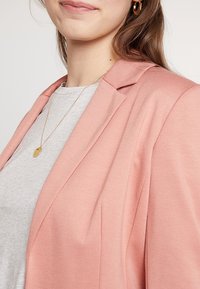 Žena oblečená v světle růžovém saku přes šedý top, se zlatým přívěškem na krku a zlatými náušnicemi ve tvaru kruhu, s dlouhými hnědými vlasy viditelnými.