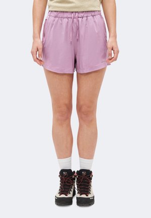 Personne portant un short lilas avec cordon, un haut beige, des chaussettes blanches et des chaussures de randonnée beiges avec semelles noires et lacets rouges, debout devant un fond uni.