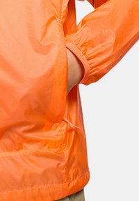 Lättvättad, klar orange jacka med elastiska cuffar, dragsko i midjan och slät textur. Sidofickor synliga.