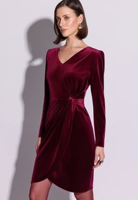 Bordowa sukienka z aksamitu z długimi rękawami, dekoltem w serek oraz marszczeniem w talii. Luźny dół opadający do połowy uda. Gładka faktura, elegancki fason.
