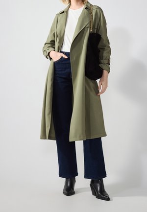 Person wearing green trench coat, white shirt, dark blue jeans, black ankle boots, holding black handbag with gold chain strap.