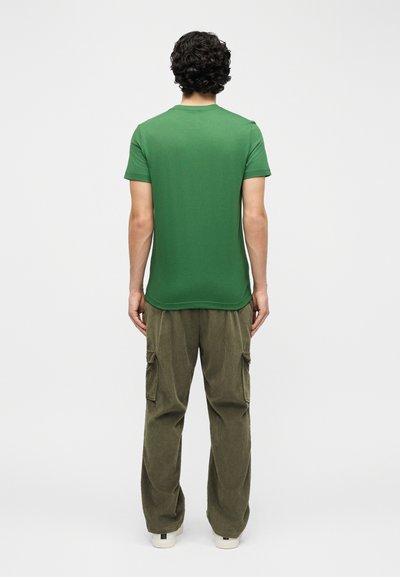 Άνδρας με σγουρά μαλλιά στέκεται με την πλάτη προς τα εμπρός, φορά πράσινο κοντομάνικο πουκάμισο, ελαιζένιο παντελόνι τύπου cargo και λευκά αθλητικά παπούτσια σε απλό φόντο.