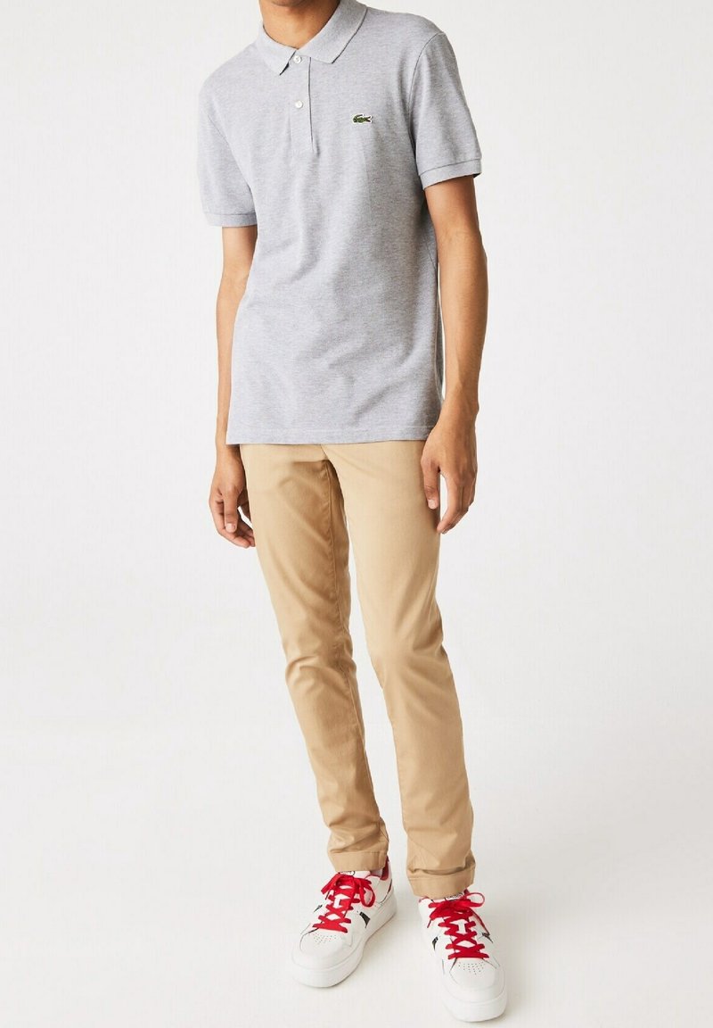 Polo gris avec un logo vert, associé à un pantalon slim beige et des baskets blanches avec des lacets rouges et des accents noirs.