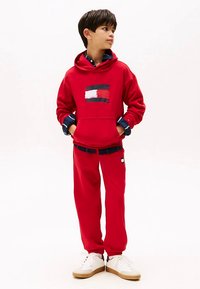 Hoodie rosso in cotone con logo, abbinato a pantaloni sportivi coordinati e sneakers bianche. Presenta un cappuccio con coulisse e polsini elasticizzati.
