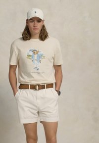 Cremefarvet t-shirt med korte ærmer og et bjørneprint, parret med hvide shorts og et brunt bælte. En hvid baseballkasket fuldender looket.