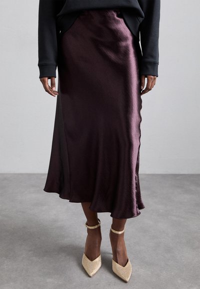 Ein tiefvioletter Satin-Midirock mit fließender Silhouette, der einen seitlichen Schlitz und einen leichten Glanz aufweist, kombiniert mit spitzen, verzierten Schuhen.