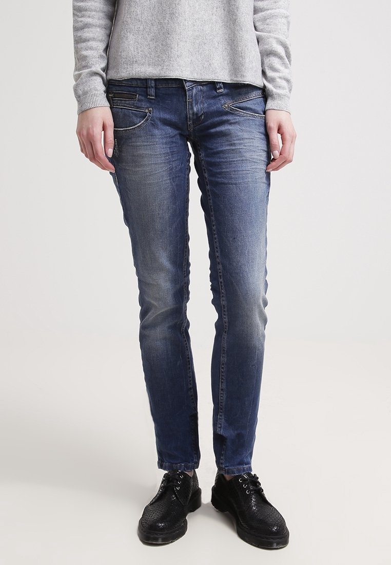 Jeans en denim bleu skinny avec un délavage, deux poches avant et des détails de couture subtils. Portés avec des chaussures noires à lacets.