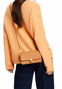 Brązowa torebka crossbody o gładkiej skórzanej fakturze, ze złotymi elementami, z klapką i długim regulowanym paskiem, noszona na pomarańczowym swetrze.