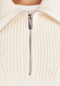 Pull de couleur crème avec un design en maille texturée, doté d'une fermeture éclair argentée au niveau du col et d'accents côtelés près de l'ourlet.