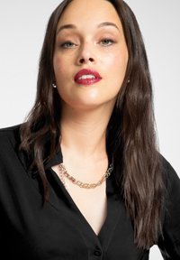 Czarna koszula na guziki z głębokim dekoltem, ozdobiona złotym łańcuszkiem. Modelka ma długie, ciemne włosy i neutralne tło.