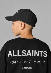 Svart keps och oversized svart t-shirt. T-shirten har stor vit text och japanska tecken. Kepsen har en böjd skärm och justerbart band.