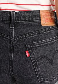 Vaqueros de mezclilla negra Levi's 501 con un parche de cuero en la cintura, costuras visibles y una etiqueta roja en el bolsillo trasero. Diseño clásico de cinco bolsillos.