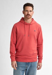 Felpa corallo realizzata in morbido tessuto, con cappuccio regolabile, polsini e orlo a coste, e un piccolo logo sul petto. Indossata con denim chiaro.