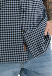 Blauwe en zwarte geruite shirt met zwarte knopen, zijzak en een bruine label op de zoom, gecombineerd met lichtblauwe spijkerbroek.