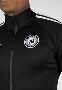 Zwarte sportjack met een hoge kraag, ritssluiting en geborduurd rond logo. Bevat witte strepen langs de schouders. Glad van textuur.
