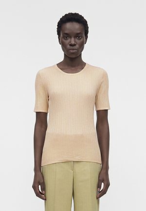 Γυναίκα με κοντά μαλλιά που φοράει μπεζ ριπ κοντομάνικο τοπ και ανοιχτό πράσινο παντελόνι ψηλόμεσο, στέκεται μπροστά σε ένα απλό φόντο.