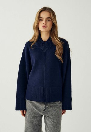Giovane donna con lunghi capelli castano chiaro che indossa un maglione blu scuro e jeans grigi, in piedi davanti a uno sfondo chiaro uniforme.