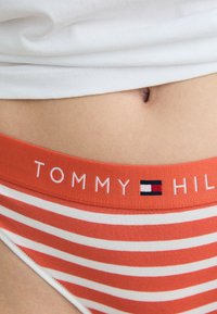 Oranssi ja valkoisiruudulliset bikini-alushousut logovyöllä, jossa on valkoisella tekstillä "TOMMY HILFIGER" ja pieni emblem. Pehmeä materiaali.