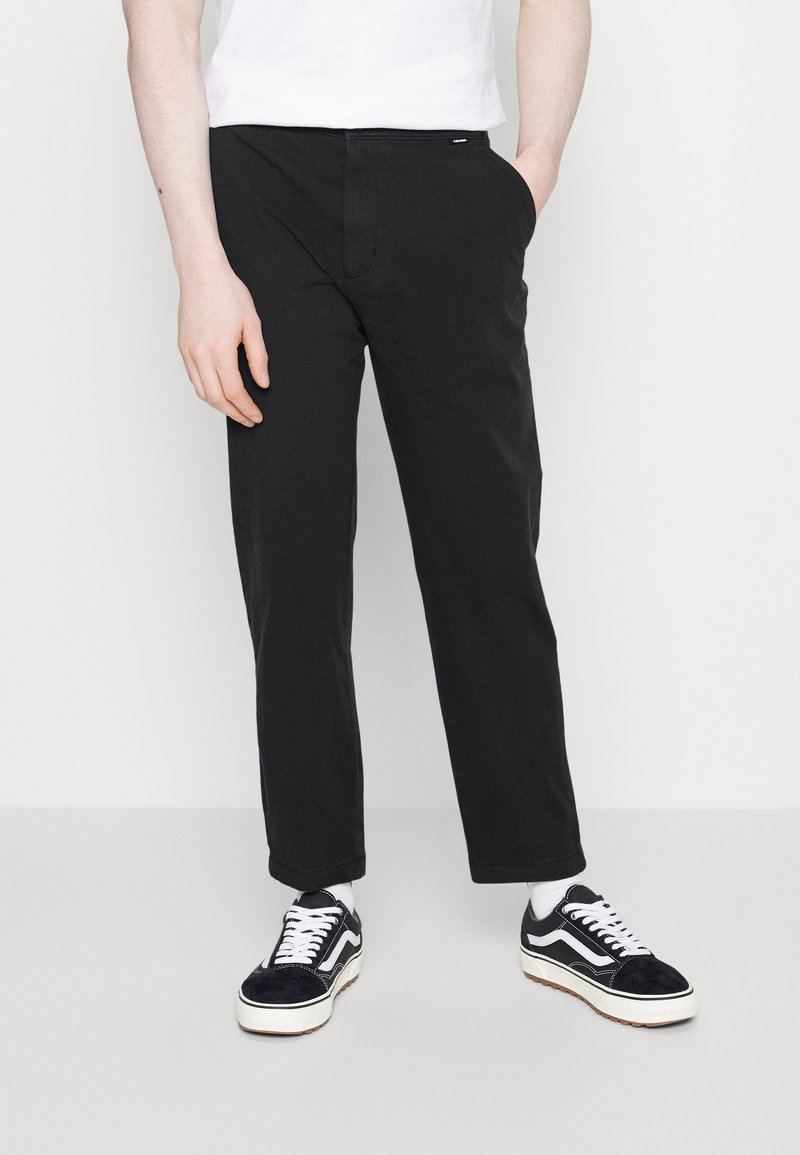 Black cotton trousers with a straight leg design, featuring side pockets and a subtle branding tag. Paired with black and white sneakers.
