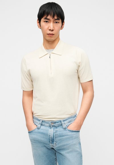 Πουκάμισο πόλο με κοντά μανίκια σε κρεμ απόχρωση, με φερμουάρ στο λαιμό, φορεμένο με ανοιχτόχρωμο τζιν, διαθέτει λεία υφή και εφαρμοστή γραμμή.