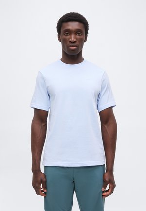 Homme debout face à l'avant portant un t-shirt bleu clair à manches courtes et un pantalon bleu sarcelle sur un fond blanc uni.