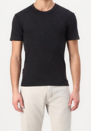 Personne portant un t-shirt noir près du corps à manches courtes et un pantalon beige clair, debout devant un fond clair uni.