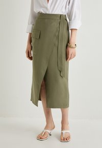 Jupe en tissu khaki enroulée avec une fente sur le côté et une poche à rabat, dotée d'une ceinture à nouer et d'une texture lisse. Portée avec des sandales blanches à bout ouvert.