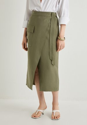 Khaki Stoffwickelrock mit einem seitlichen Schlitz und einer aufgesetzten Tasche, ausgestattet mit einem Bindegürtel und glatter Textur. Tragen zu weißen Sandalen mit offenem Zehenbereich.