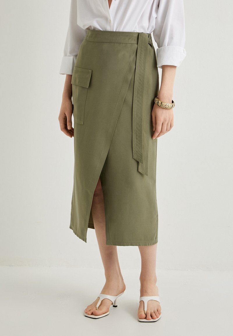 Jupe en tissu khaki enroulée avec une fente sur le côté et une poche à rabat, dotée d'une ceinture à nouer et d'une texture lisse. Portée avec des sandales blanches à bout ouvert.