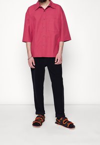 Roze kortemouwen overhemd met een zak, gecombineerd met donkere broeken en sandaal met oranje en gele banden.