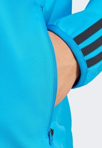 Giacca sportiva blu con tasca con zip, texture liscia e strisce nere che accentuano i polsini per un tocco di dettaglio.