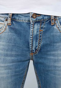 Jeans de denim azul con costuras naranjas, con una cintura de altura media, diseño de cinco bolsillos y un cierre de botón en la parte frontal.