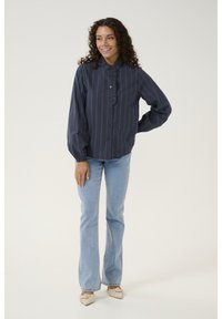 Marineblauwe blouse met knopen en verticale witte strepen, met een geruisde kraag en lange pofmouwen. Gecombineerd met lichtblauwe flare jeans en beige schoenen.