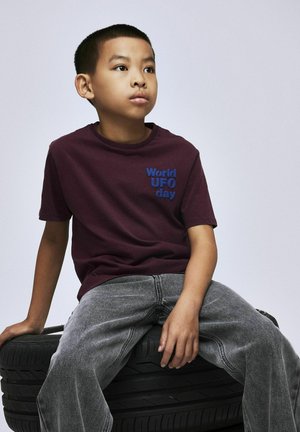 Bordeauxrotes Baumwoll-T-Shirt mit blauer Schrift "Welt-UFO-Tag", kombiniert mit grauen Jeans. Das Kind sitzt auf einem schwarzen Reifen vor einem neutralen Hintergrund.