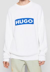Biały sweter dziany o prążkowanej fakturze, z niebieską prostokątną naszywką z logo "HUGO" w białych literach na wysokości klatki piersiowej.