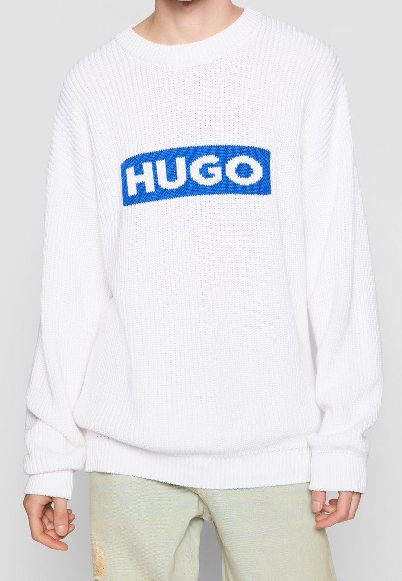 Biały sweter dziany o prążkowanej fakturze, z niebieską prostokątną naszywką z logo "HUGO" w białych literach na wysokości klatki piersiowej.