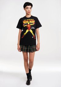 Schwarzes Baumwoll-T-Shirt mit einem bunten Iron Man-Graphic auf der Vorderseite, kombiniert mit einem grünen karierter plissierter Mini-Rock und schwarzen Schuhen.