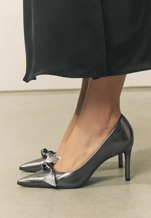 Metalen grijze hoge hakken met een spitse neus en een strikaccent aan de voorkant. Strakke textuur met een glanzende afwerking.