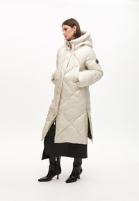 Lång, puffig jacka i ljusbeige, med huva, diamantquiltning, sidofickor och svart logotypmärke. Bärs över en svart outfit och stövlar.