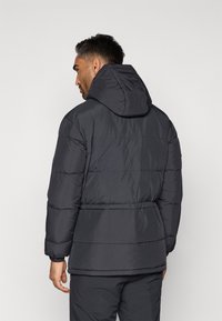 Homem com cabelo curto e escuro, vestindo uma jaqueta acolchoada preta com capuz e calças pretas, em pé e de costas, contra um fundo simples.