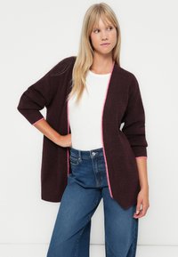 Cardigan bourgogne avec des bordures roses, à l'avant ouvert et aux manches trois-quarts. Porté par-dessus une chemise blanche et un jean bleu taille haute. Texture douce.