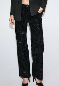 Pantalones negros de pernera ancha con tejido texturizado que presenta un patrón floral y lentejuelas. Combinados con una chaqueta negra ajustada y tacones de punta.