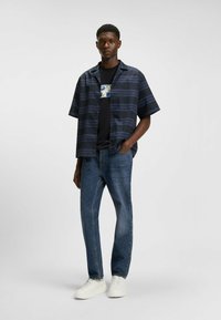 Short-sleeve navy striped shirt, black graphic t-shirt underneath, blue denim jeans, and white sneakers, standing against a neutral background.