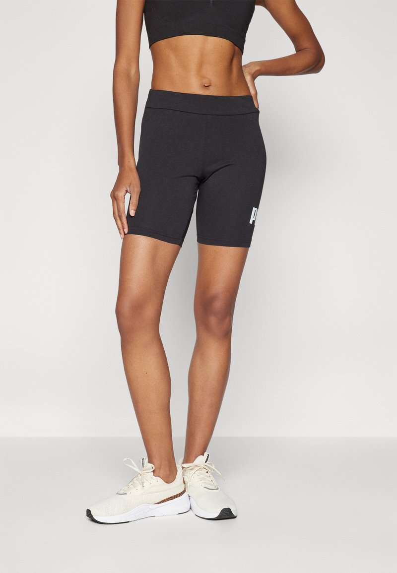 Černé elastické šortky s vysokým pasem, se světlým logem na boku, doplněné světle barevnými teniskami.