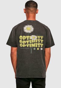 Schwarzes Baumwoll-T-Shirt mit großen weißen Gänseblümchen mit gelben Mittelpäckchen und dem Text "OPTIMIST" in verschiedenen Größen und Farben auf dem Rücken.