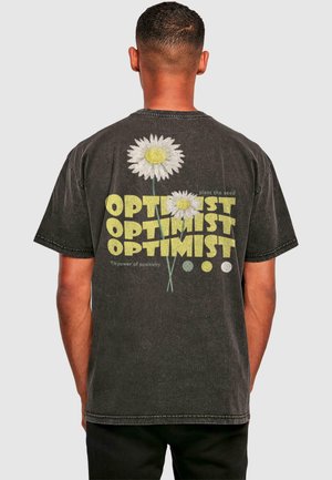 Sort bomulds T-shirt med store hvide margueritter med gule midterpunkter og teksten "OPTIMIST" i forskellige størrelser og farver på ryggen.