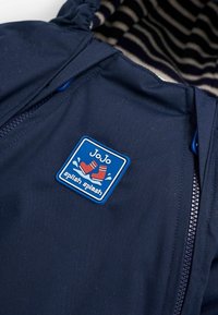 Marine waterdichte jas met een gestreepte binnenkant, een rits aan de voorkant en een blauwe rechthoekige patch met rode laarzen en de tekst "Jo Jo splish splash".