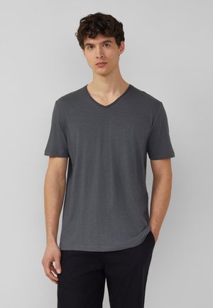 Mann steht vor einfachem Hintergrund und trägt ein dunkelgraues V-Ausschnitt-Kurzarm-T-Shirt und schwarze Hose, mit einer Hand in der Tasche.